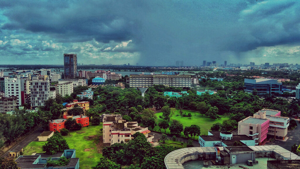 The city of Kolkata on a cloudy day. In the foreground are buildings and green parks. In the distance, a large body of rain is falling over part of the city.