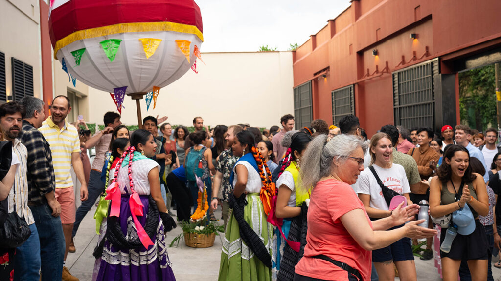 Several dozen people socialize in an open-air courtyard between two buildings. A large sphere covered in white fabric and adorned with colorful flags stands on a pole above some of the people.