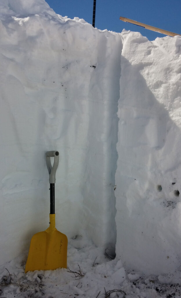 A yellow shovel stands at the bottom of a 6-foot snow pit.
