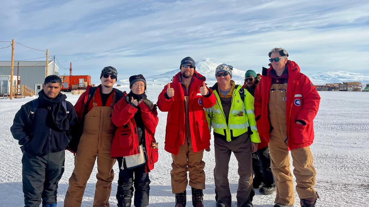 Seven people pose for a photo on an icy field with a mountain in the background on a sunny day. The man in the middle gives a thumbs-up.