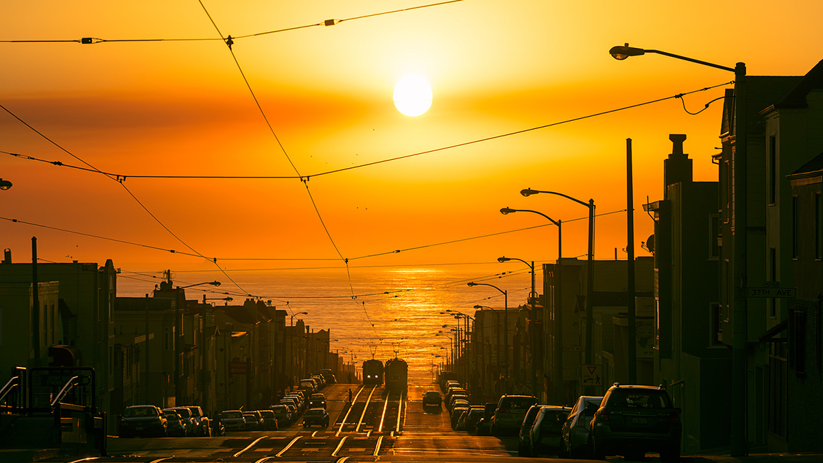 A shot from atop a hill in San Francisco looks down at the ocean and an approaching cable car. The sun over the ocean is creating an orange glow,andmostof the buildings and cars are seen in silhouette.