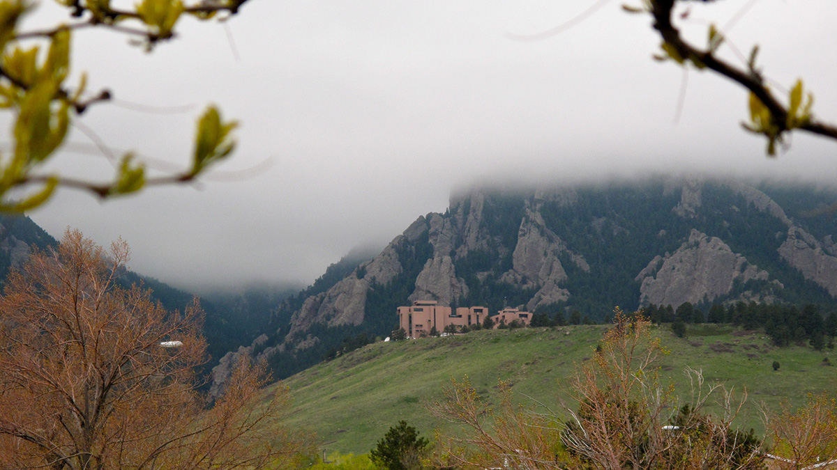 A building on a hillside has a foggy sky and mountains just behind it and green rolling hills in front of it. The image is framed by branches in the foreground.
