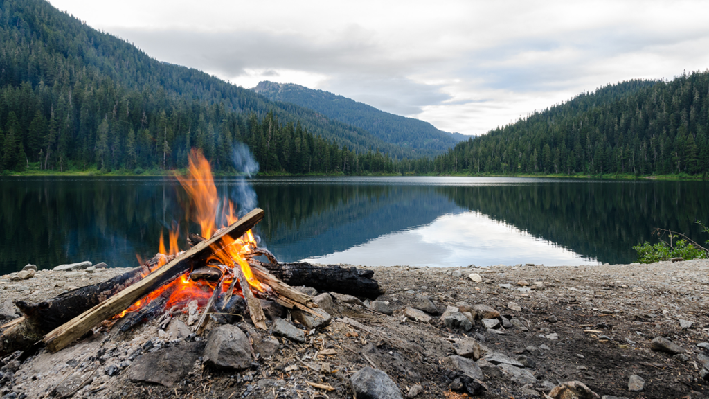 A small camp fire overlooking a peaceful lake with mountains in the background.