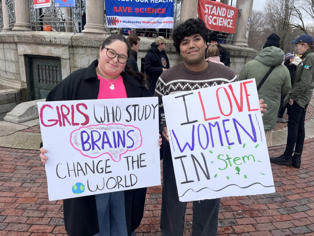 Two people hold signs at a rally. One sign, held by a woman wearing a pink shirt and black sweater, says “Girls who study brains change the world.” The other sign, held by a man wearing a brown sweater, says “I love women in STEM.”
