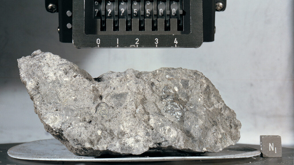 A gray, speckled rock sits on a metal plate. Above the rock is a sign showing the numerical label of the rock sample, 7225500.