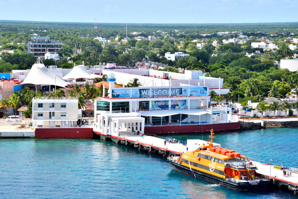 Aerial view overlooking an ocean cruise terminal. An orange ferry is docked along a long pier extending from the terminal building, which has a large “Welcome” sign atop it.