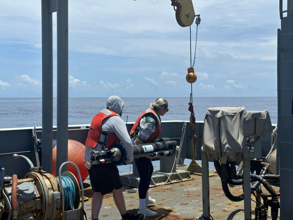 Two people wearing orange life preservers and standing on the deck of a ship hold a torpedo-shaped float.