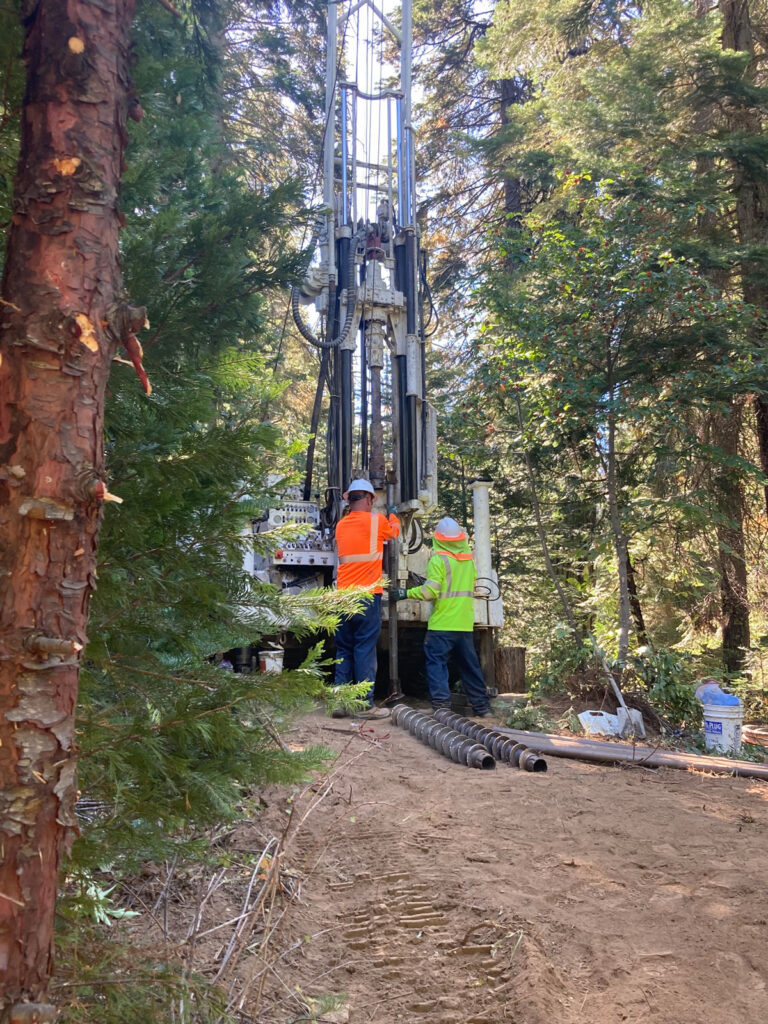 Two people stand by a large drilling rig situated on a dirt track among tall trees in a forest.