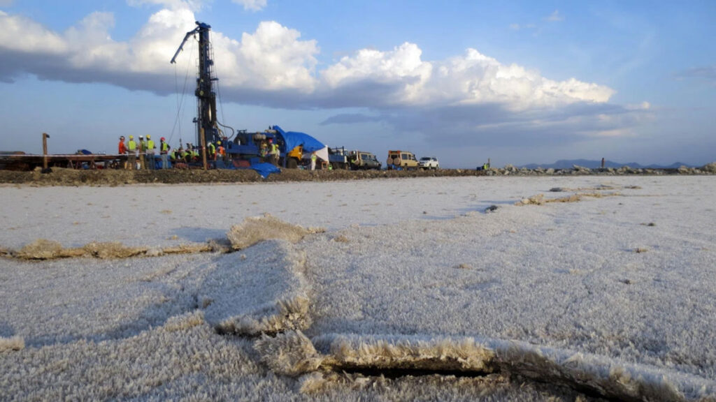 Ground-level view looking over a flat expanse of land covered in a crust of crystalline salt, with a group of people standing around a tall drilling rig in the distance.