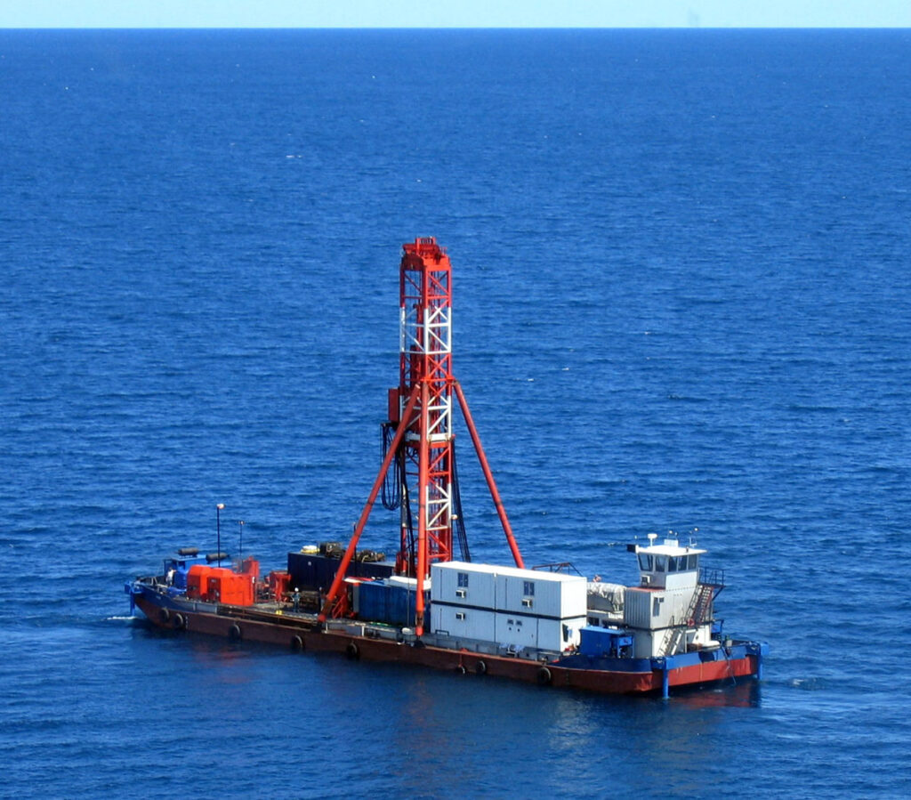 Low-angle aerial view of a barge equipped with a tall, red-and-white-painted drilling rig sitting in an expanse of blue water.