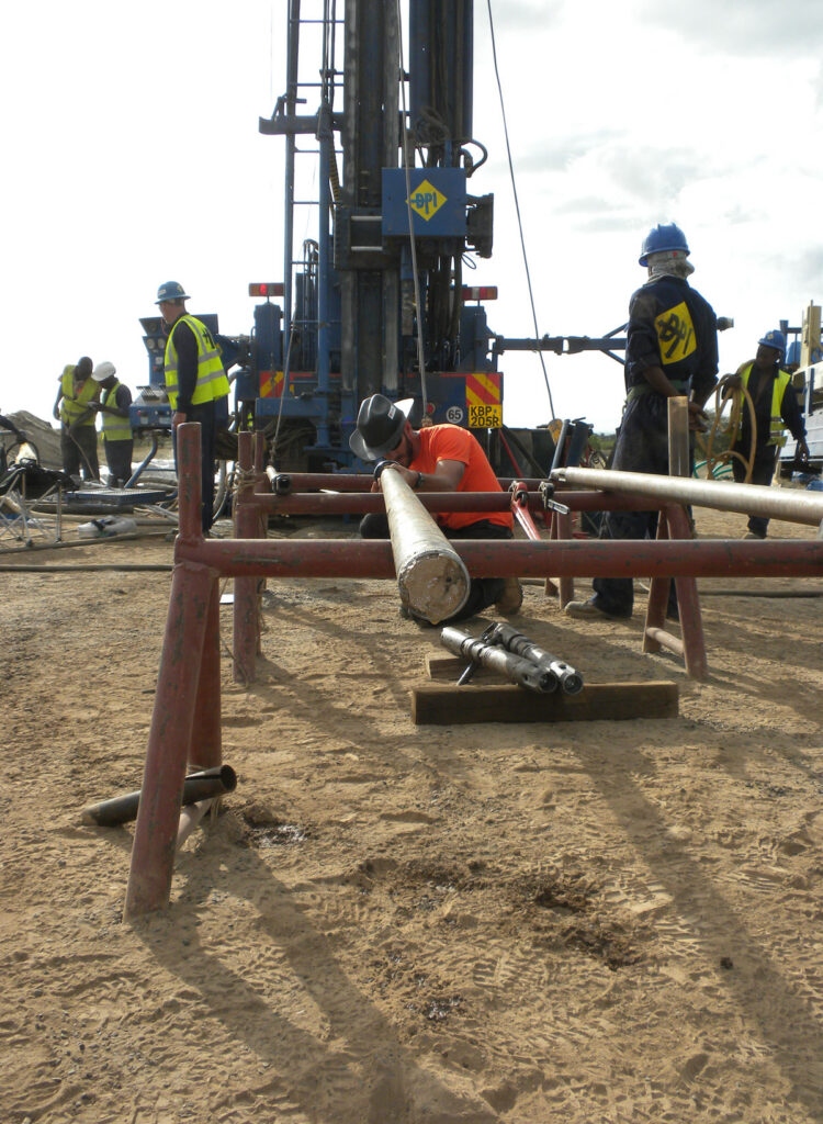 View looking down the length of a narrow sedimentary rock core sitting on a metal framework. In the background, several people stand around a large drilling rig and a person wearing a cowboy hat in a bright orange shirt looks down the core from the other end.