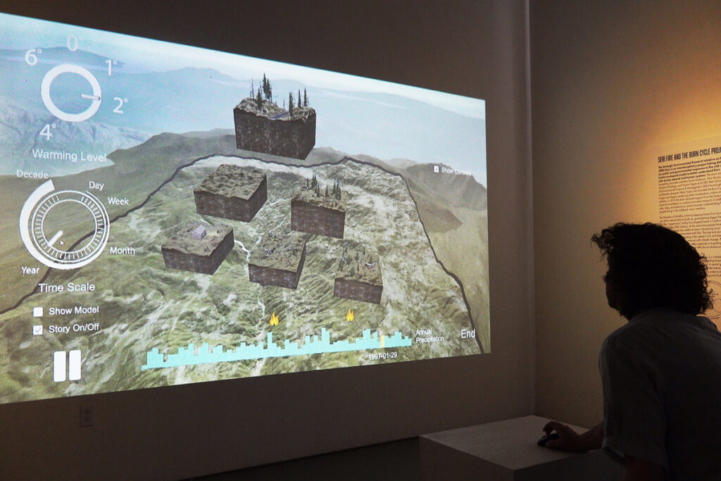 A silhouetted museumgoer uses a computer mouse to make selections in an interactive video exhibit, which depicts a hillside watershed and is projected onto a blank wall.