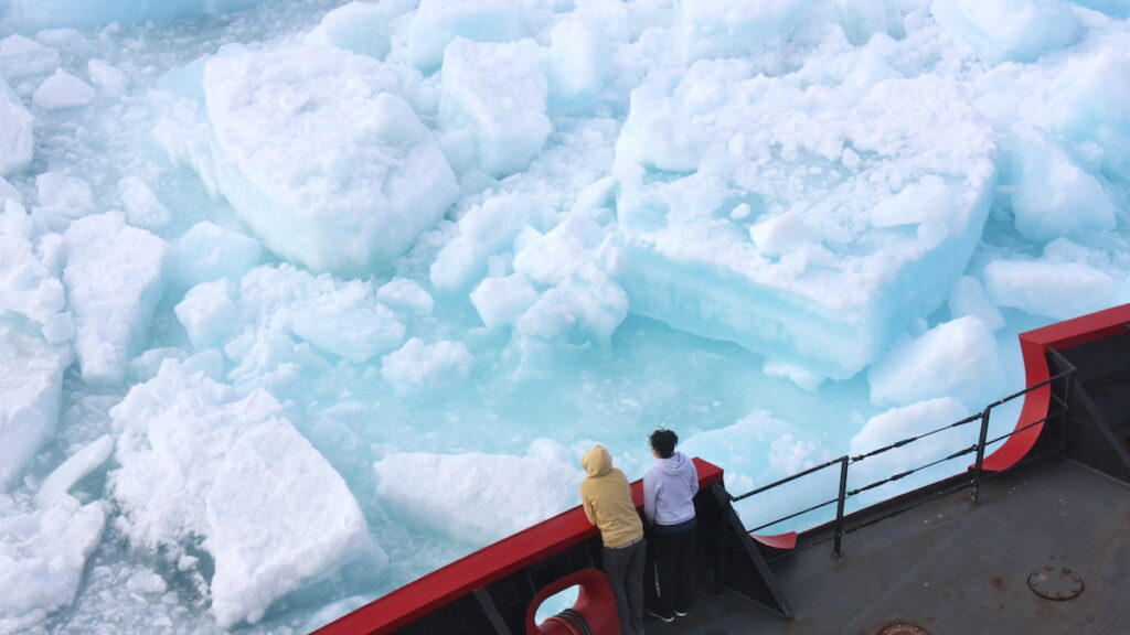 Two people on the deck of a large ship look over the edge of the ship at large chunks of floating ice.