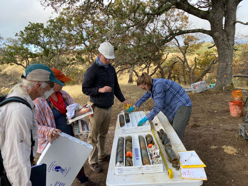 A researcher handles sandstone core samples placed in a white box atop a white picnic table outside while three other people look on.