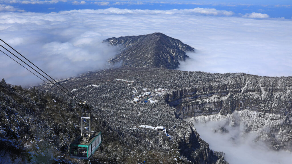 A mountain of gray rock rises above clouds, and a gondola is descending toward the mountain.