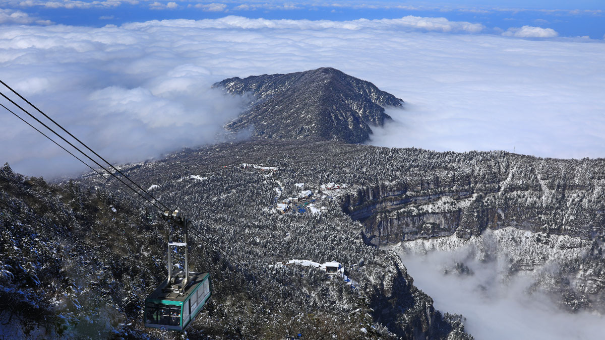 A mountain of gray rock rises above clouds, and a gondola is descending toward the mountain.