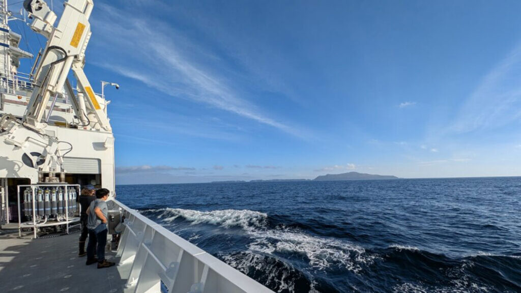 Two people look out at the ocean over the starboard side of a research ship out at sea.