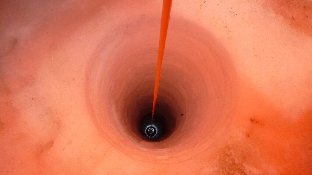 A hole drilled through a glacier extends off into the darkness. A camera is being lowered into the hole. The photo is bathed in red light.