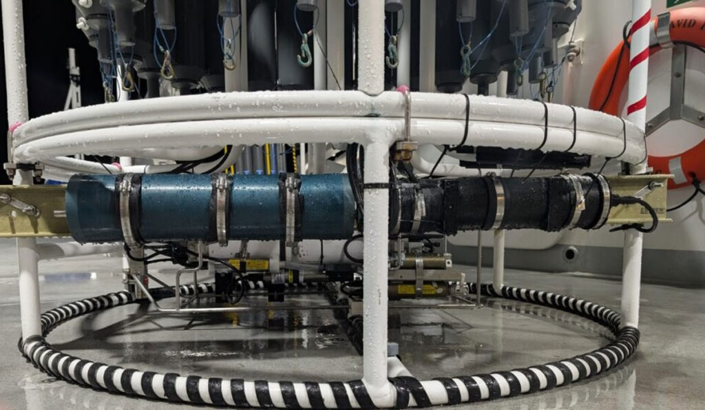 The base of a circular sampling rosette dripping with water sits on a deck of a research ship. Two cylindrical spectrophotometers, one blue and on black, can be seen attached to the frame of the rosette, constructed of cylindrical white tubing.