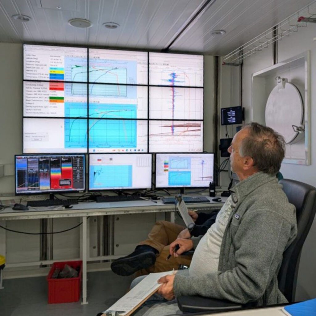 A person sits in a desk chair holding a clipboard and pen and looking at a display of scientific data projected across nine wall-mounted monitors, as well as at other information shown on three monitors sitting on a desk.