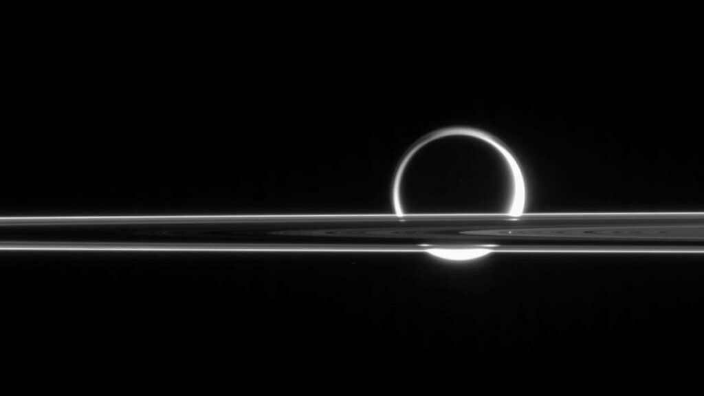 The Sun shines through the edges of Titan’s atmosphere, making it look like a ring of fire in black and white. In the foreground, Saturn’s concentric rings are brightly lit.