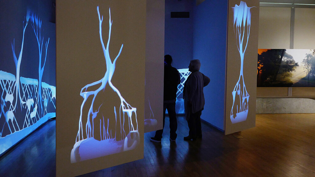 Two people stand in a darkened space in an art gallery amid stylized images of trees and flowing water projected in shades of light to dark blue onto walls and hanging screens.