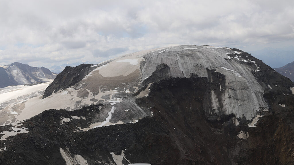 The summit of a glacier is largely dark soil, showing how the ice is melting.