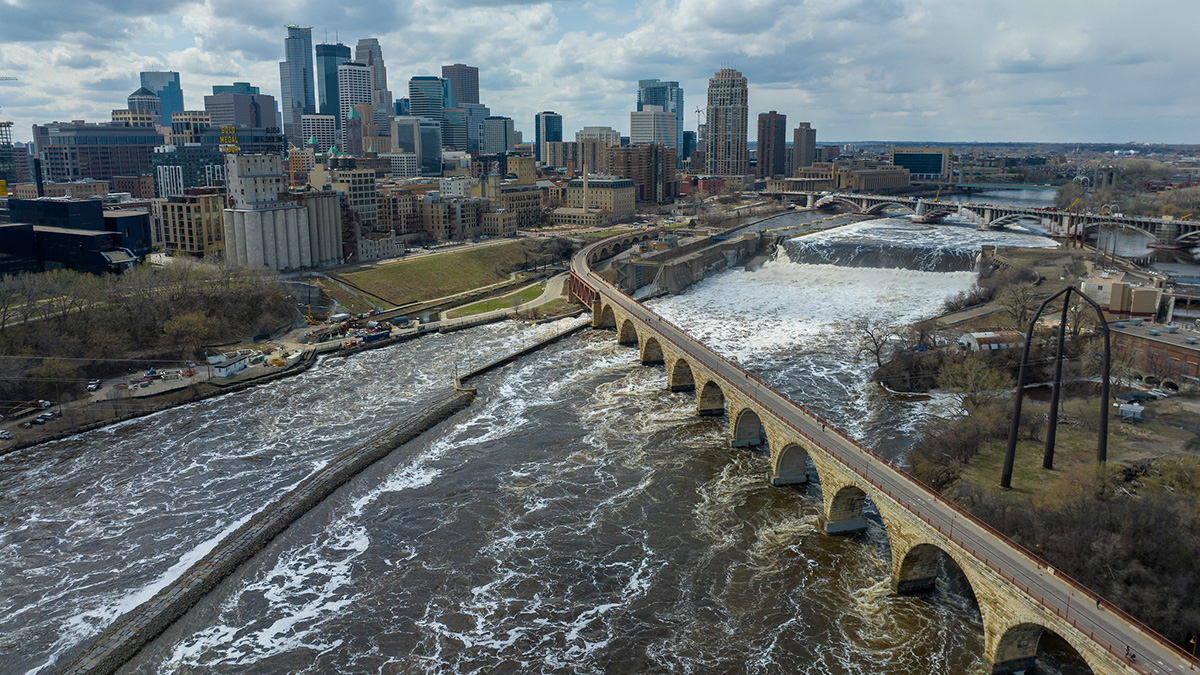 A bridge crosses a river beneath a relatively short waterfall. A city skyline is on the other side of the river.