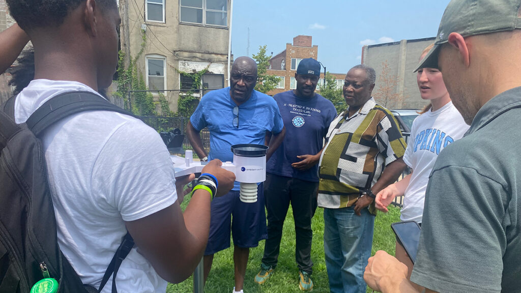 Six adults stand around a tripod that holds a personal weather station device. They are standing on grass, and several buildings are visible not far behind them.