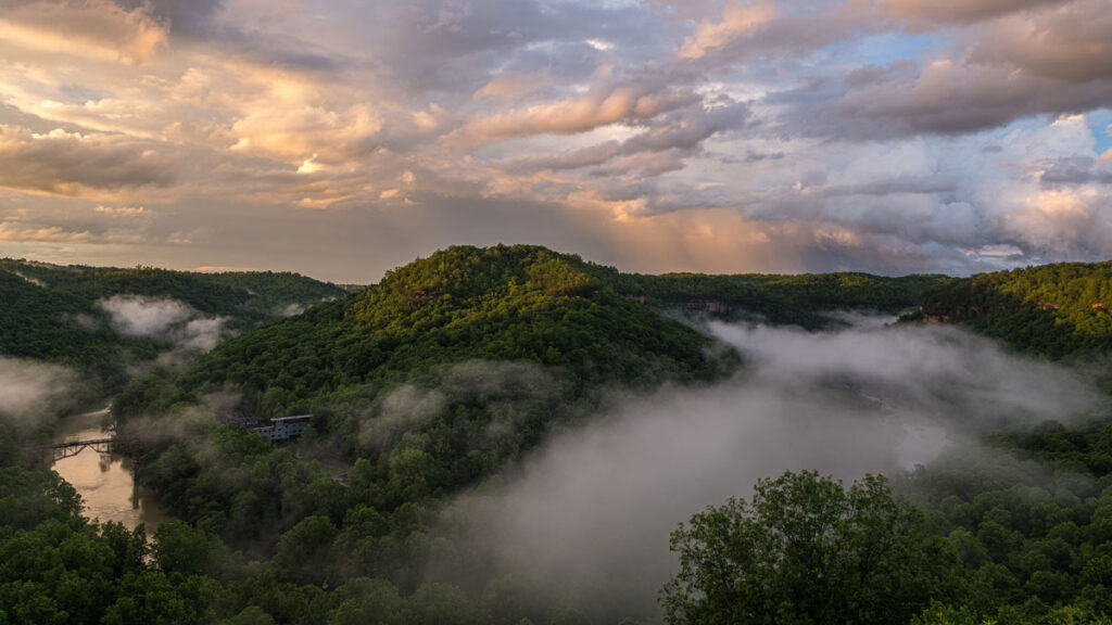 A foggy mountain scene at sunset. In the right-hand corner, a railroad leading to a small building can be seen.