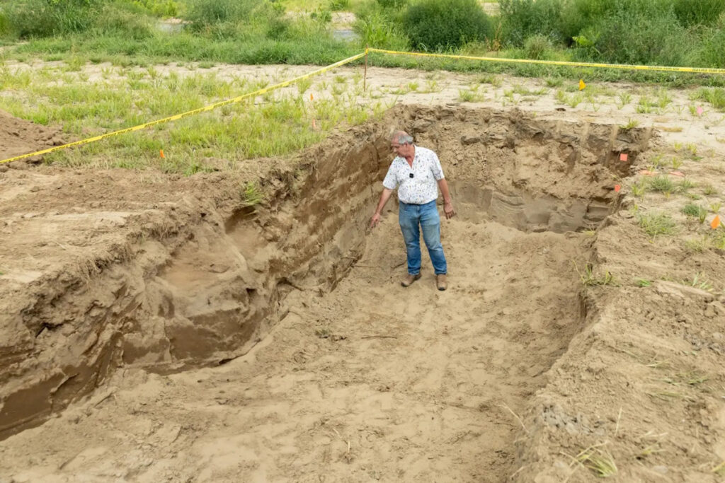 A man stands in a rectangular dirt pit, about the same height as himself, which is surrounded by yellow caution tape.