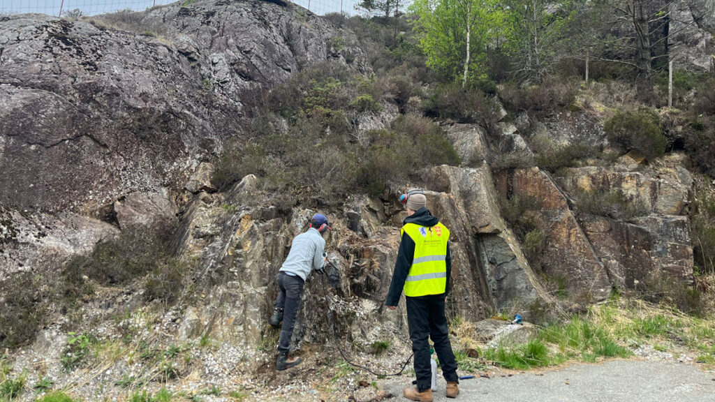 Two people, one wearing a yellow vest and one in a gray long-sleeved shirt, look up at a rock face.