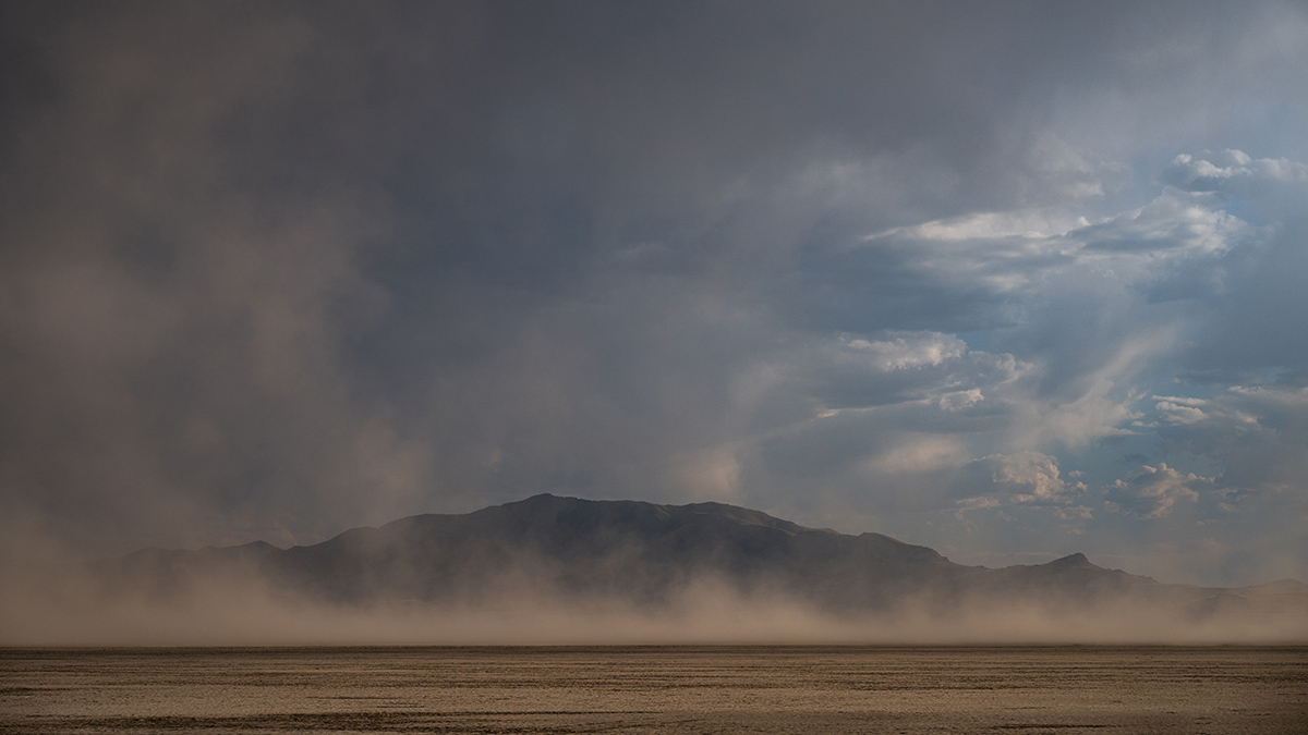Dust clouds rise from a dry, flat expanse of lake bed in front of mountains and a partly cloudy sky in the background.
