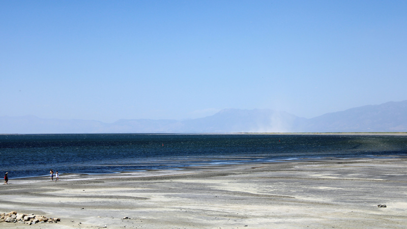 Three people stand under clear skies near the shoreline of the Great Salt Lake on a wide, flat expanse of dry lake bed. A small plume of dust rises in the distance on the far side of the lake, in front of a line of silhouetted mountains.