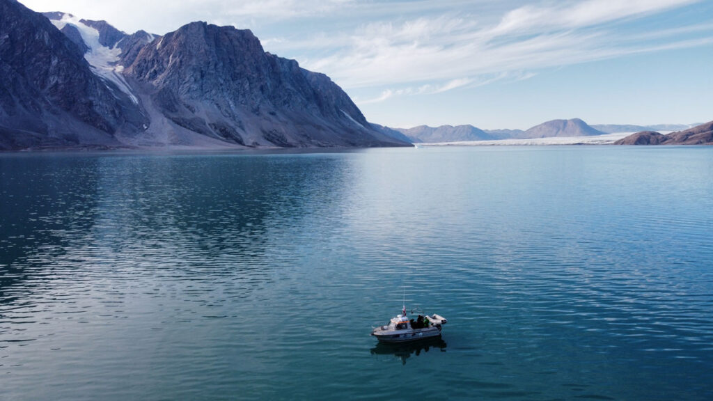 Aerial view of a small research boat sitting in blue-green water in front of glaciated mountains that rise steeply from the shoreline.