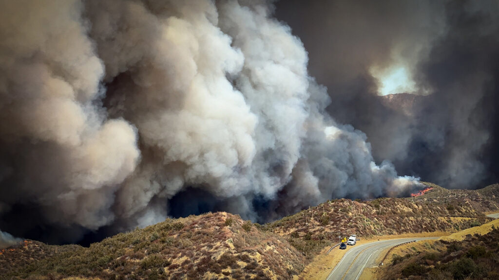 Large plumes of smoke rise from a hillside. Two vehicles on a roadside are dwarfed by the smoke cloud.