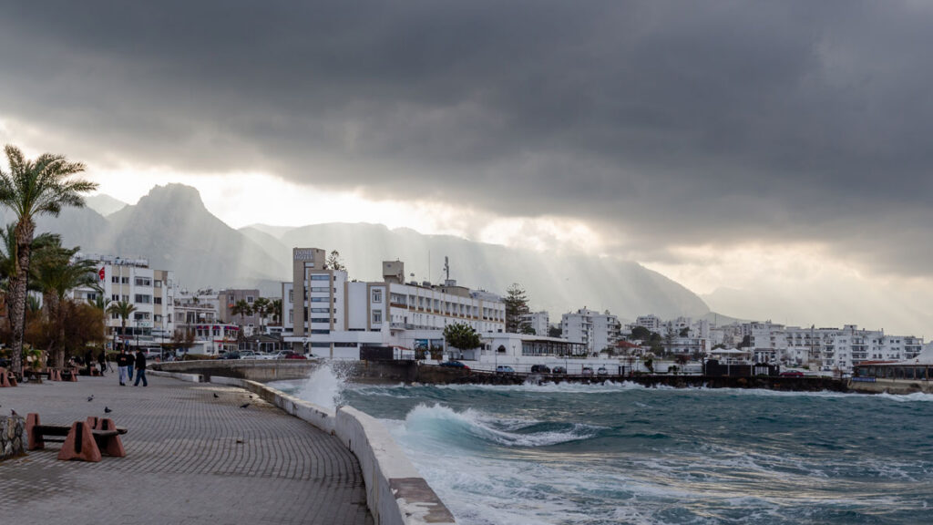 A dense urban development is seen on a shoreline. Ominous clouds herald the onset of a storm, and waves lap against the shore.