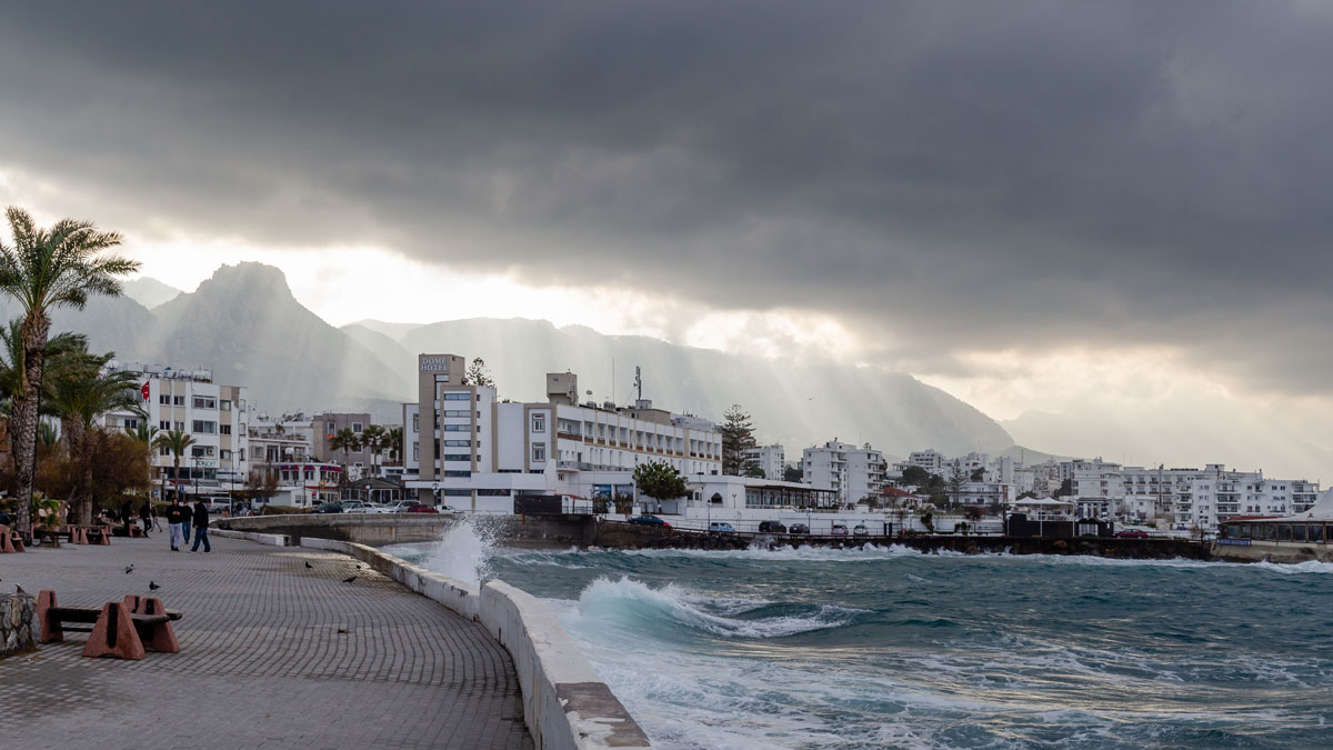 A dense urban development is seen on a shoreline. Ominous clouds herald the onset of a storm, and waves lap against the shore.