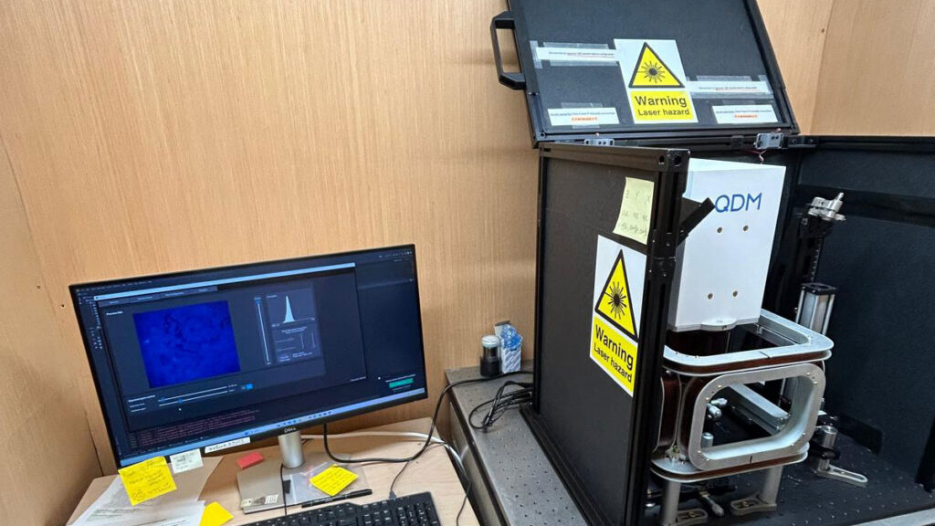 A computer and keyboard on a desk sit next to a complex microscope that says “QDM” on the top.