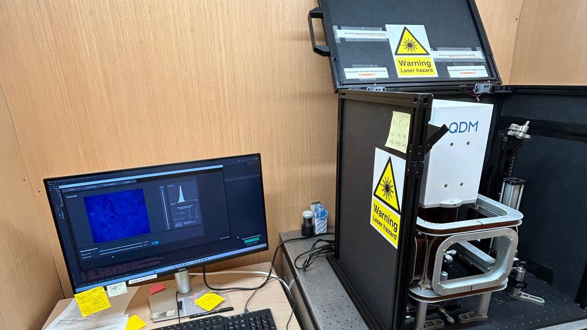 A computer and keyboard on a desk sit next to a complex microscope that says “QDM” on the top.