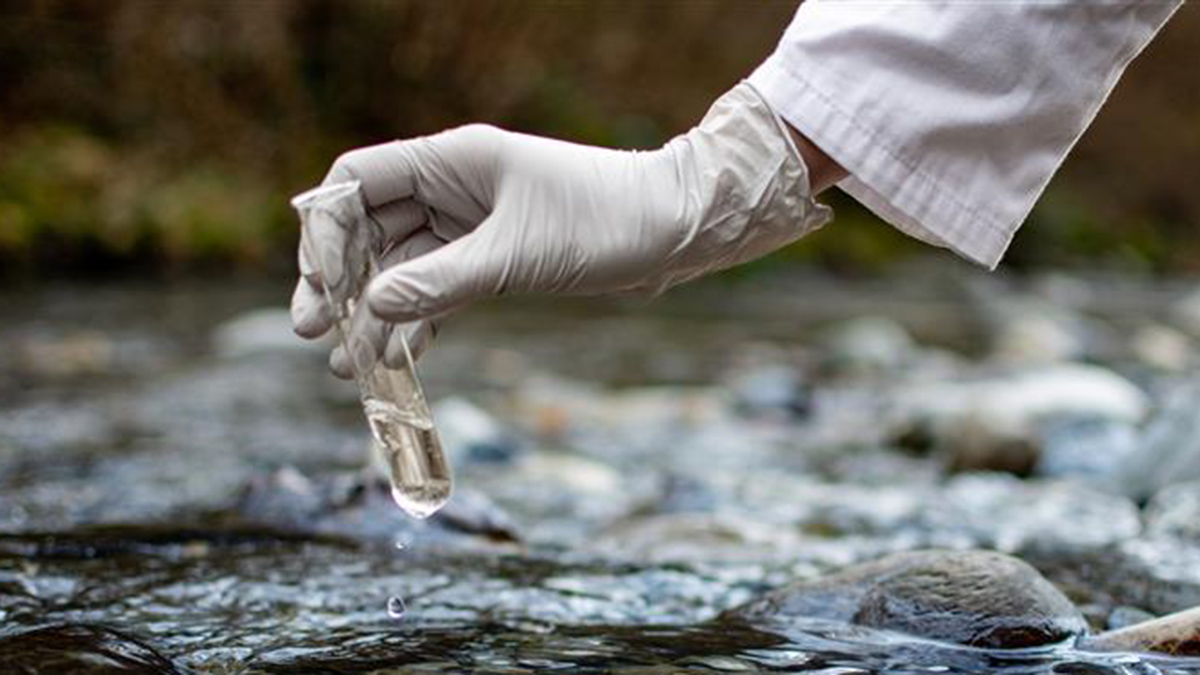 A person’s right arm extends into the frame from the right over a running stream. The gloved hand holds a test tube partially filled with water that’s just been collected; a partial droplet of water is collecting at the bottom of the tube.