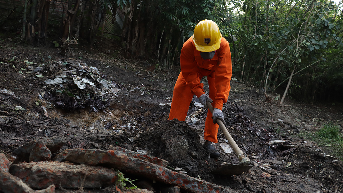 A person in an orange jumpsuit and a yellow hard hat takes a tool similar to a hoe to the dark soil they are standing on.