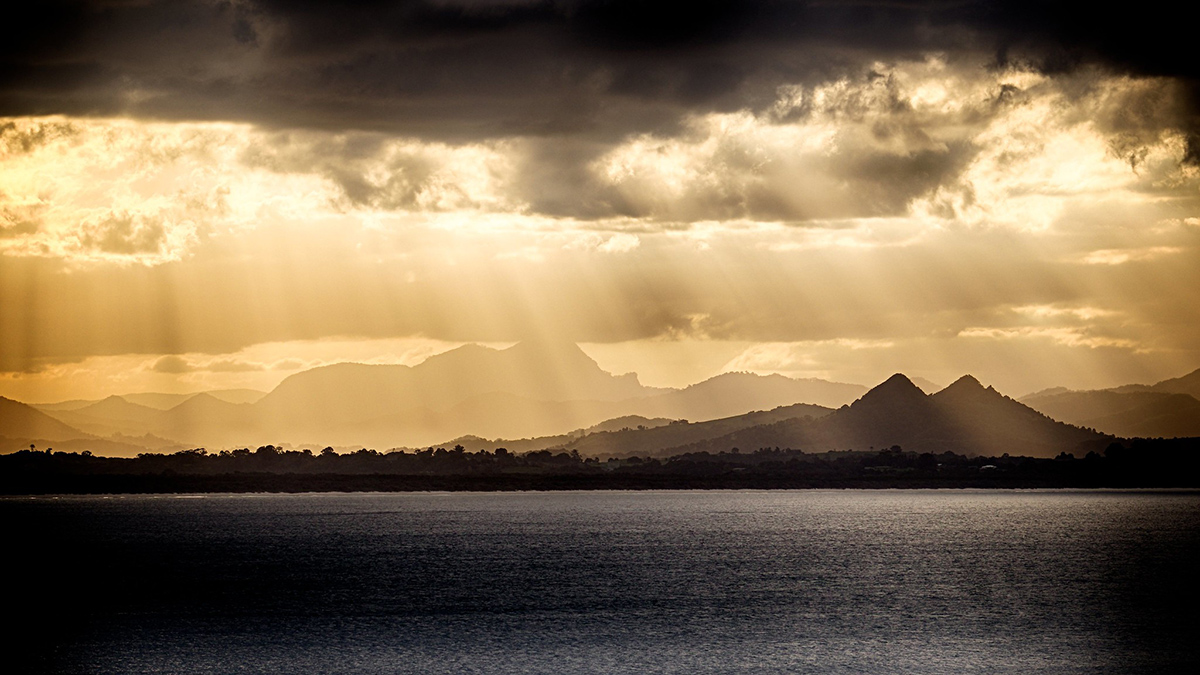Yellow light shines through a layer of clouds onto the ocean below. A volcano is visible, in silhouette, in the distance.