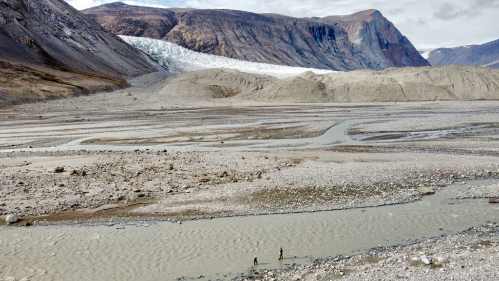Aerial view looking across braided river streams and channel bars toward mountains, a glacier, and hills of glacial deposits in the distance. In the foreground, two researchers stand in the gray, sediment-filled waters of one of the streams.