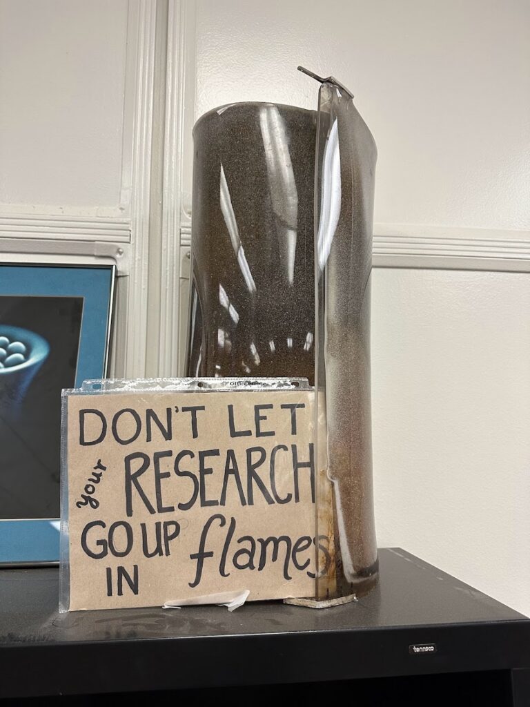 A piece of charred plastic rests on a surface. A sign leaning against the plastic reads, “Don’t let your research go up in flames.”