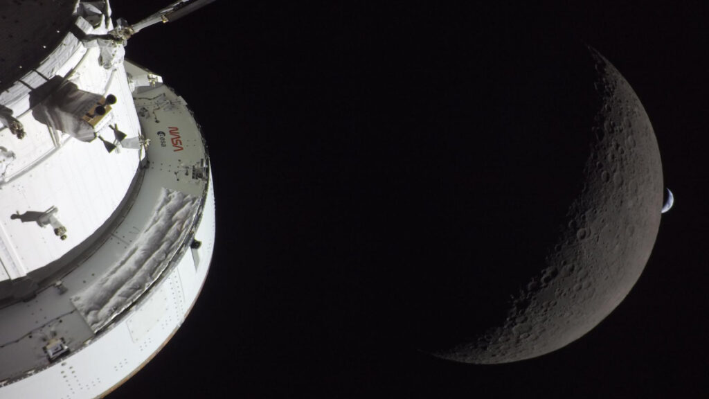 A photo of the Orion spacecraft in front of a crescent of the farside of the Moon, which is in front of a crescent of the Earth in the distance