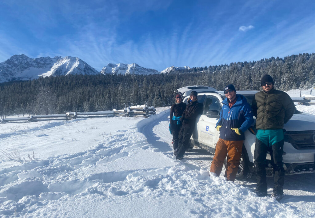 Four people lean against a white sport utility vehicle in a snowy field, with forest, mountains, and a cloud-streaked blue sky behind them.