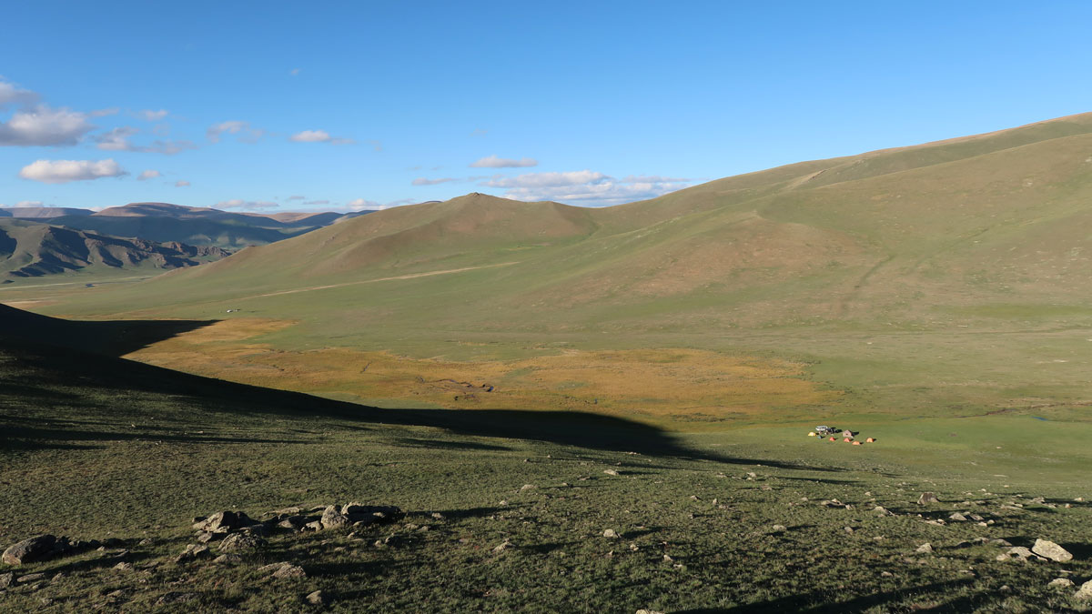 The gentle green slopes of a mountain range with a small field camp nestled at the base.
