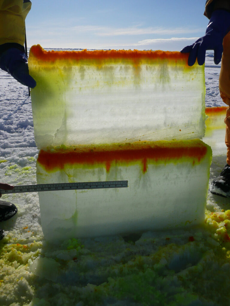 Two blocks of ice, about half the height of an adult man, are stacked on top of each other. Both are red along the top, and the red dye is moving down through the ice in some points.