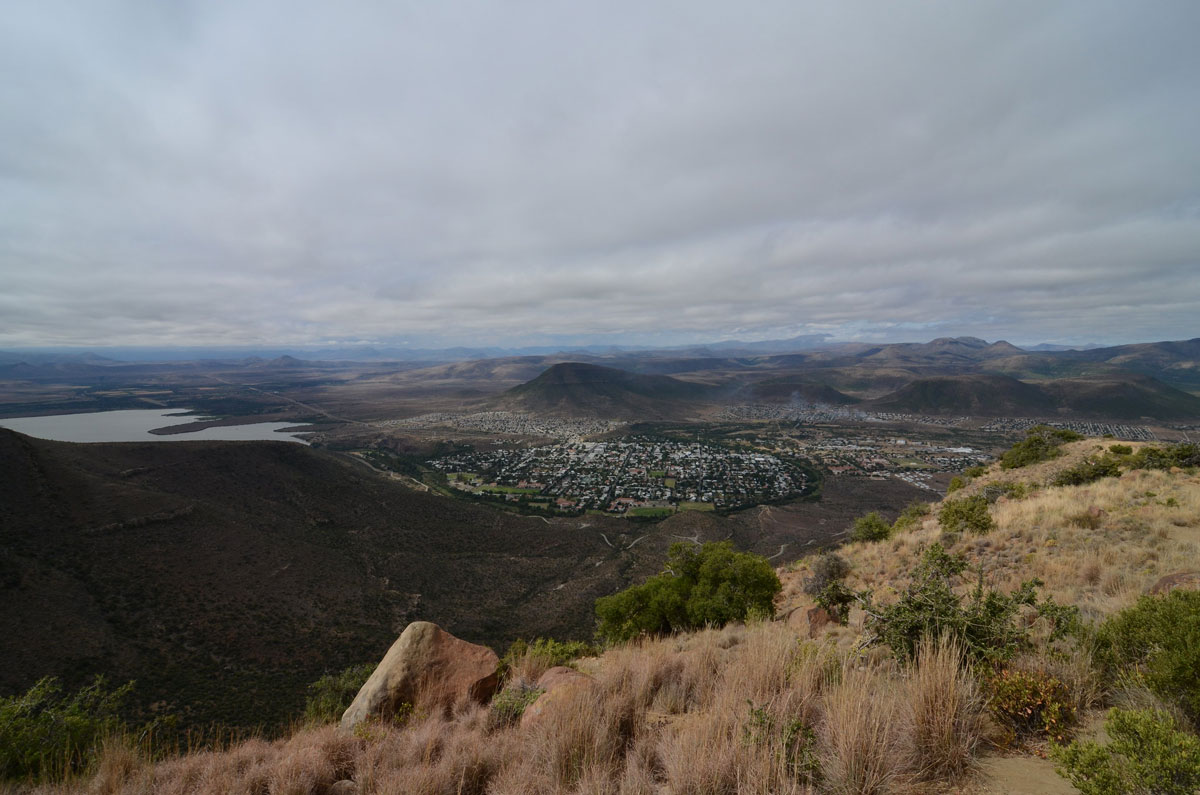 A photo taken from on a hill shows a populated valley surrounded by brown mountains on a cloudy day.